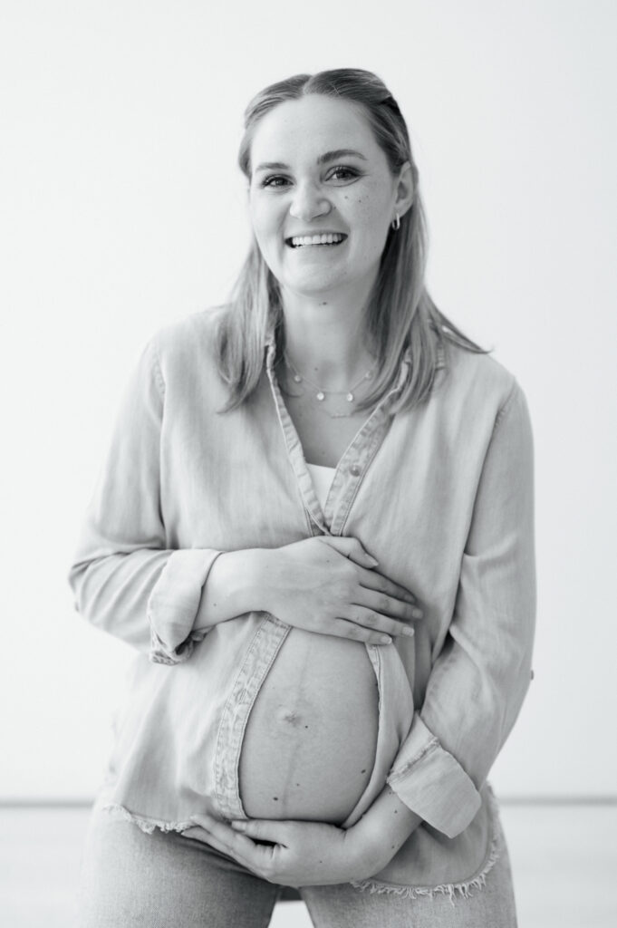 expecting mom embraces baby bump and smiles at camera during studio maternity photos