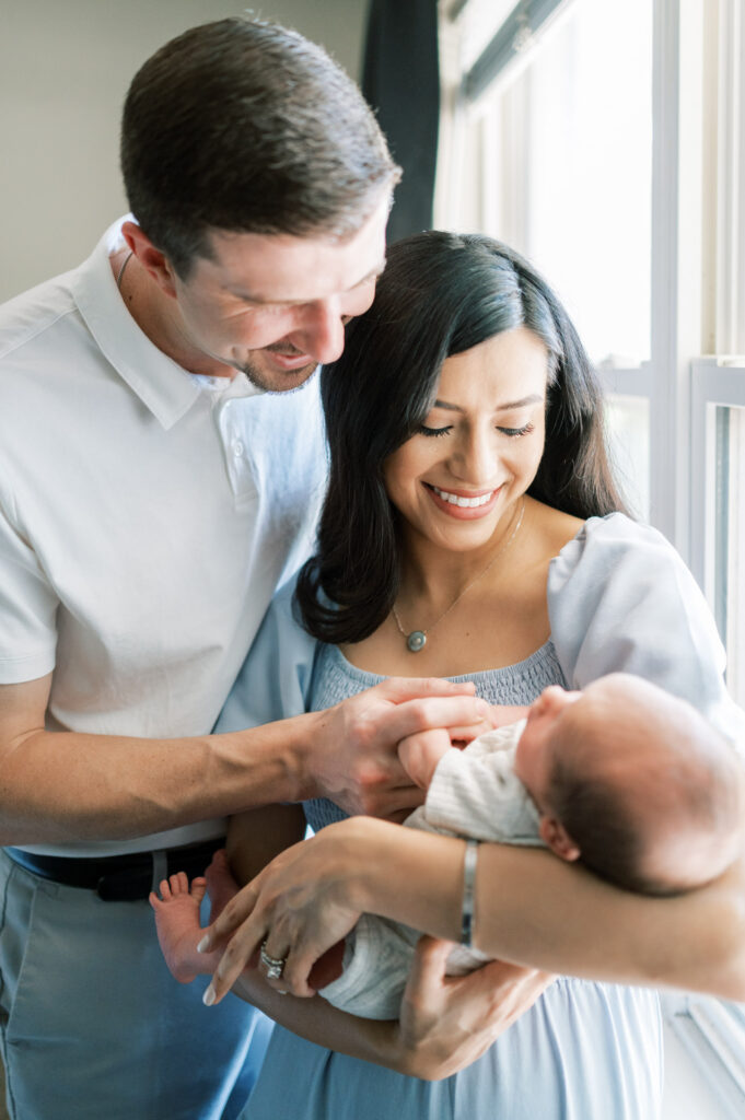mom and dad stare adoringly at baby newborn near bedroom window