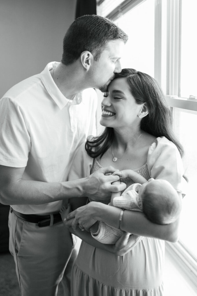 dad leans in and kisses new mom while she holds baby newborn
