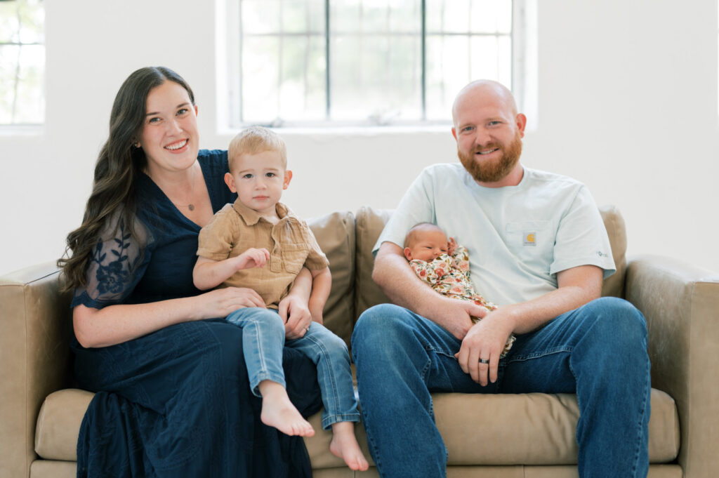 a family of four with a newborn sits on a couch in a bright studio space