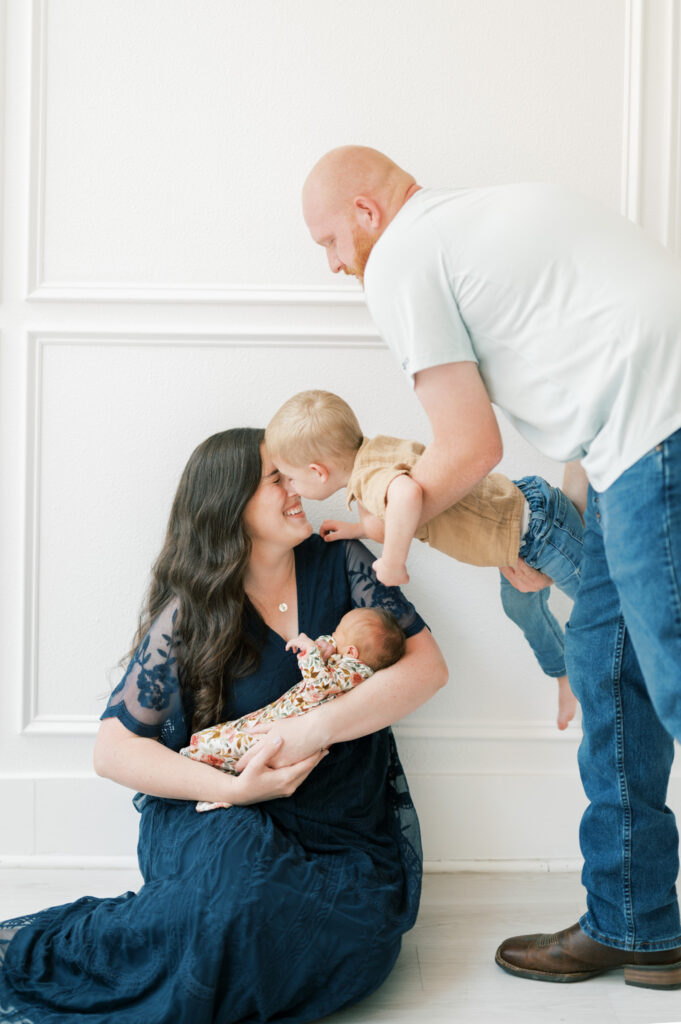 a dad airplanes his son towards mom to give her a kiss while she holds a baby