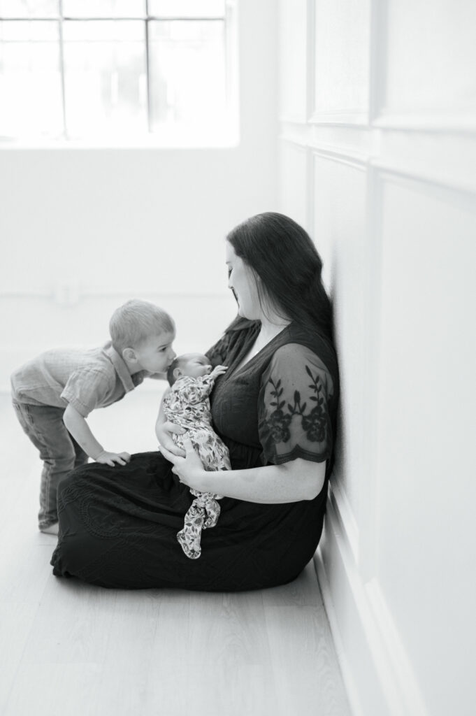 a little boy kisses his newborn sister's head in a studio while mom holds baby