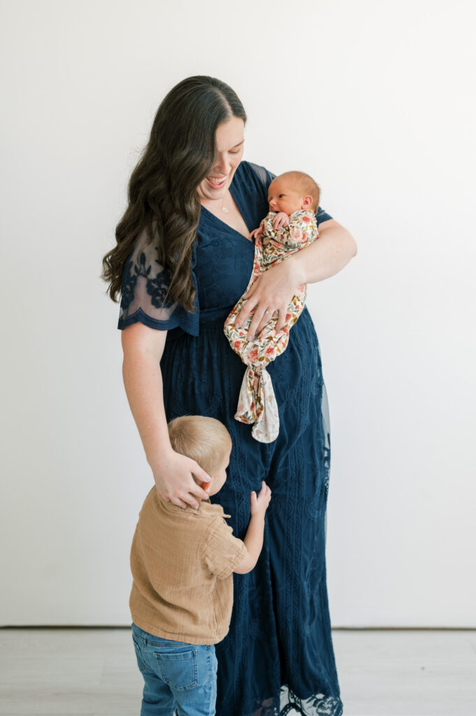 a little boy hugs his mom's leg while she holds a newborn in a studio