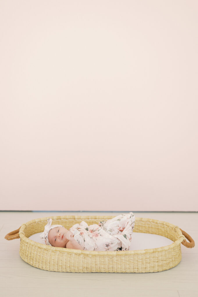 baby girl lies in basket in front of pink backdrop