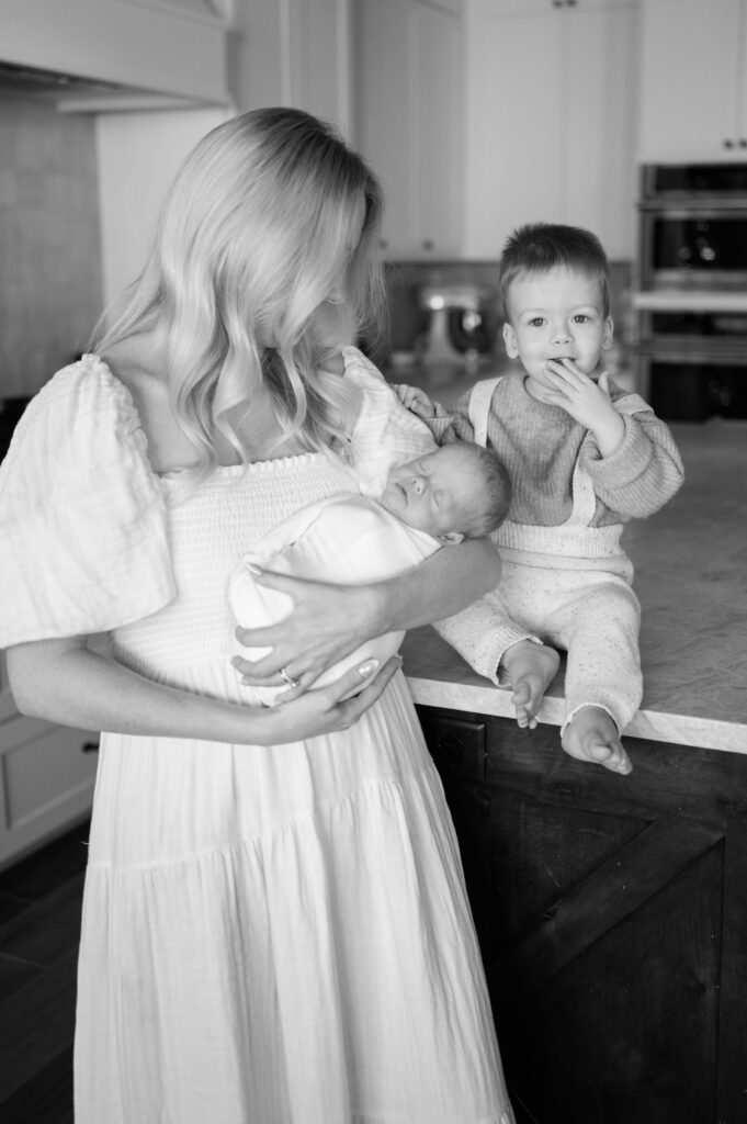 mom stands in kitchen with newborn baby and big brother