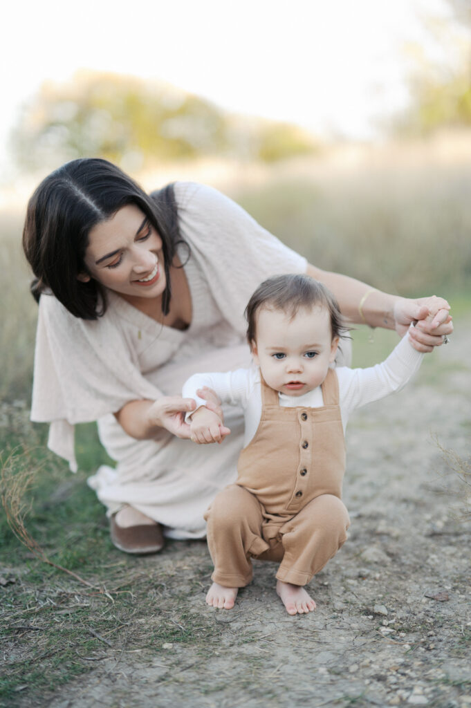 mom holds hands of one year old while he stands on a gravel path