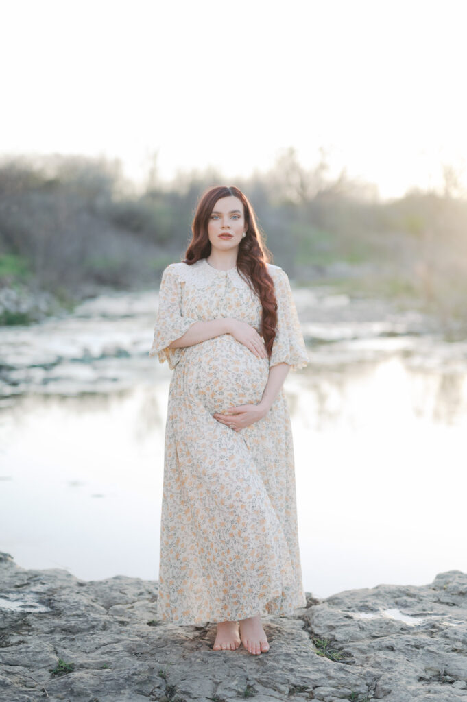 mom embraces baby bump standing in front of creek, looking at camera