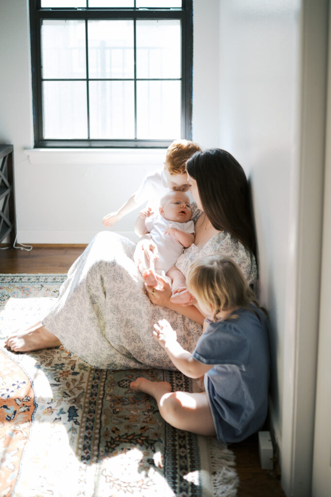 mom sits on floor in sunlight with newborn baby and two toddlers