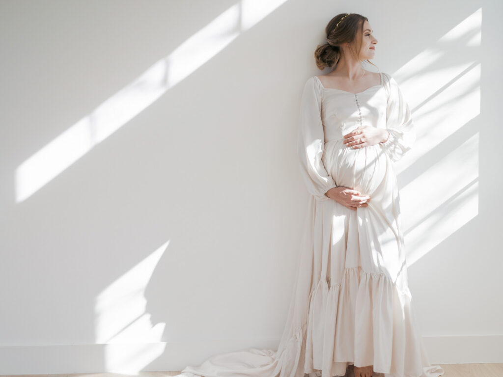 expecting mom leans against a wall during maternity photos with light shining throuh