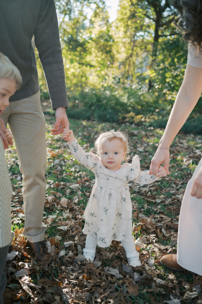 toddler girl looks at camera while walking and holding parents hands
