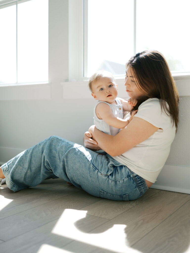 mom holds baby boy sitting by bright window while sun shines through