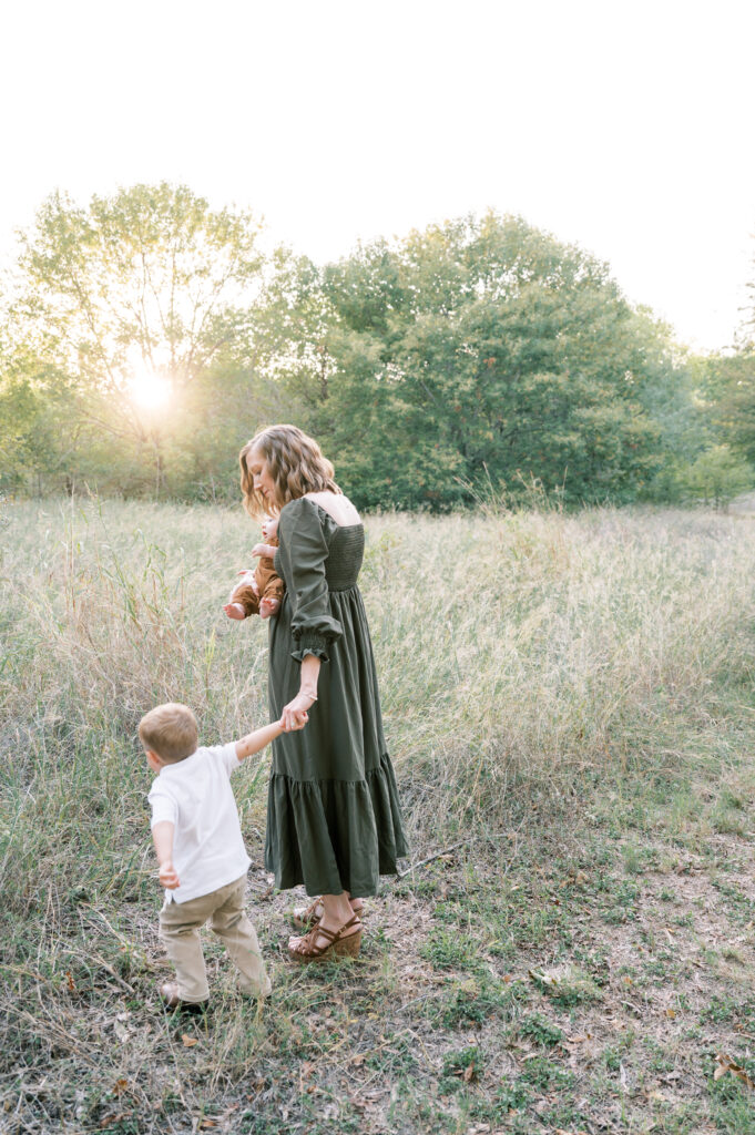 mom carries baby and holds toddler's hand as they walk into a field