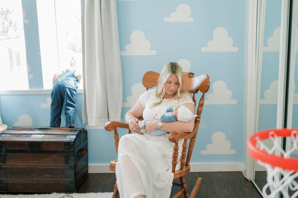 mom rocks newborn baby while toddler looks out bedroom window during in-home newborn session
