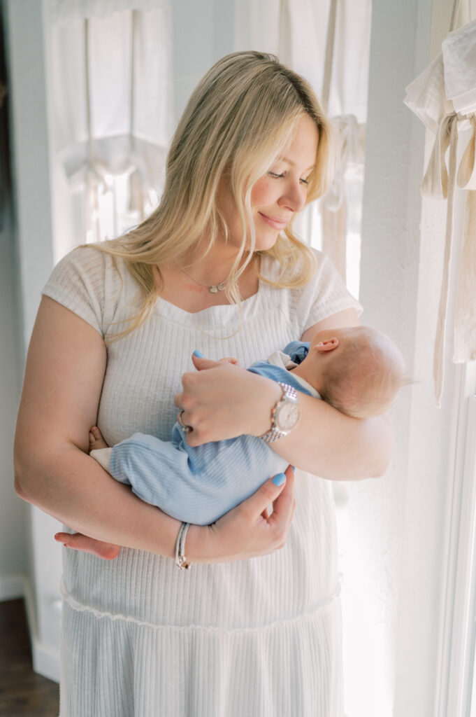 mom looks lovingly at newborn while next to window