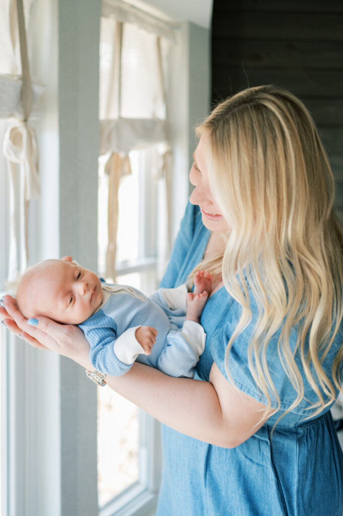 mom holds baby newborn next to window and smiles during in-home newborn session