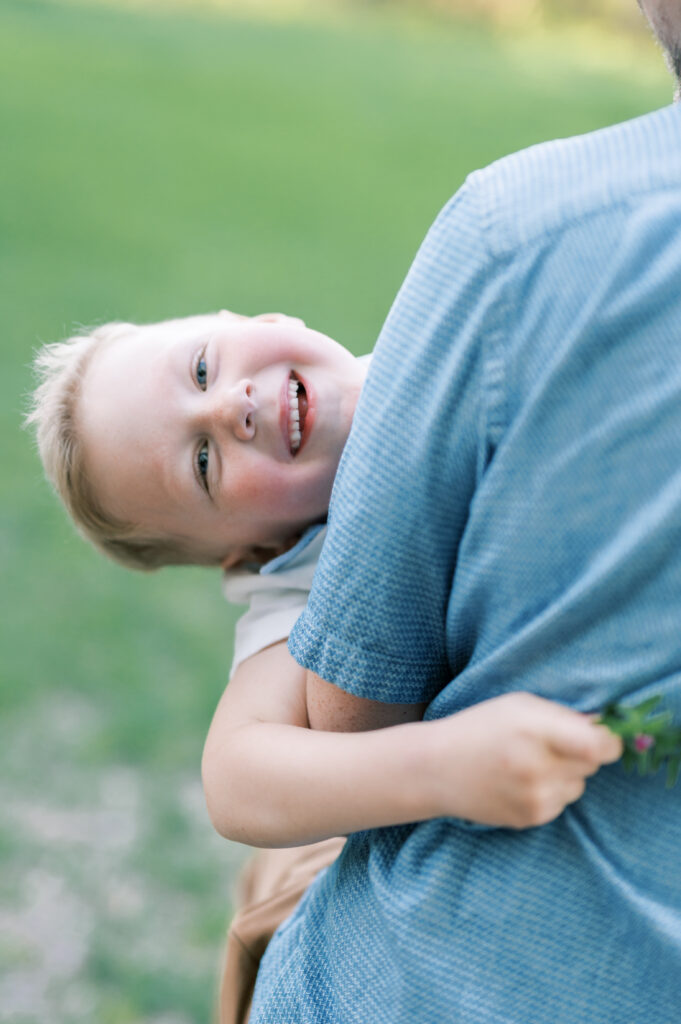 a dad carries a boy while he looks back at the camera and smiles