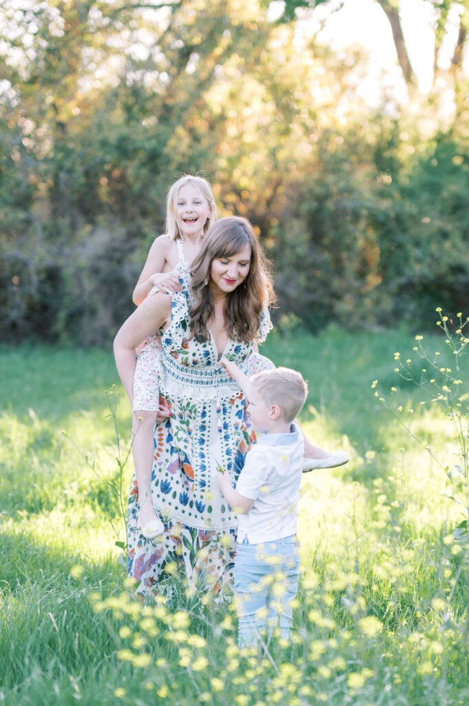 mom carries daughter in a field while son hands her a flower