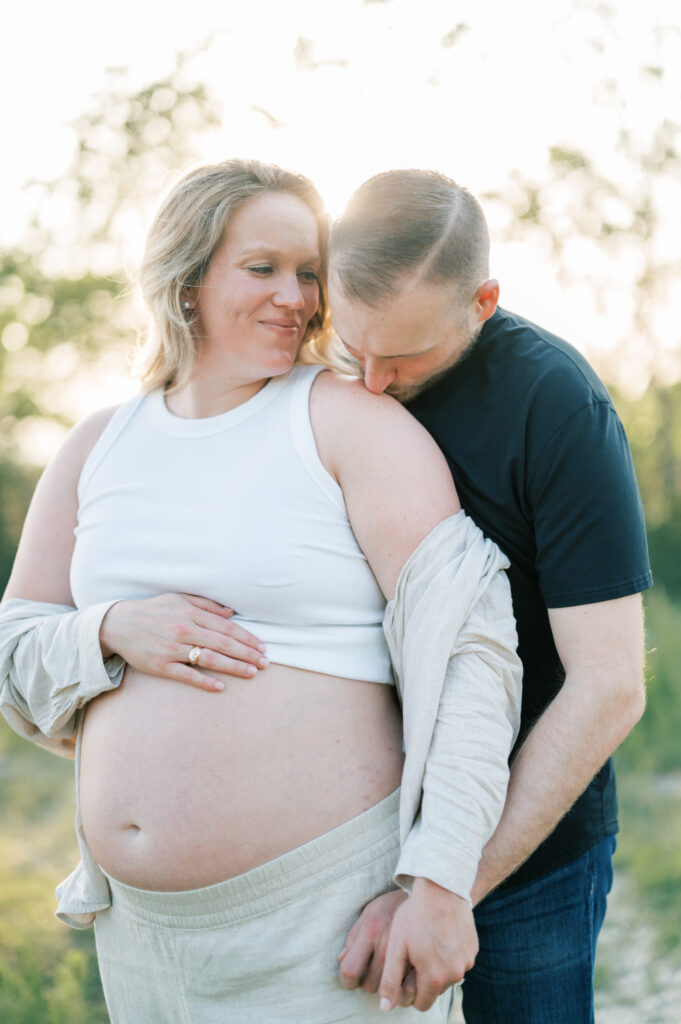expecting dad sweetly kisses wife's shoulder while sunlight reflects onto them during maternity session