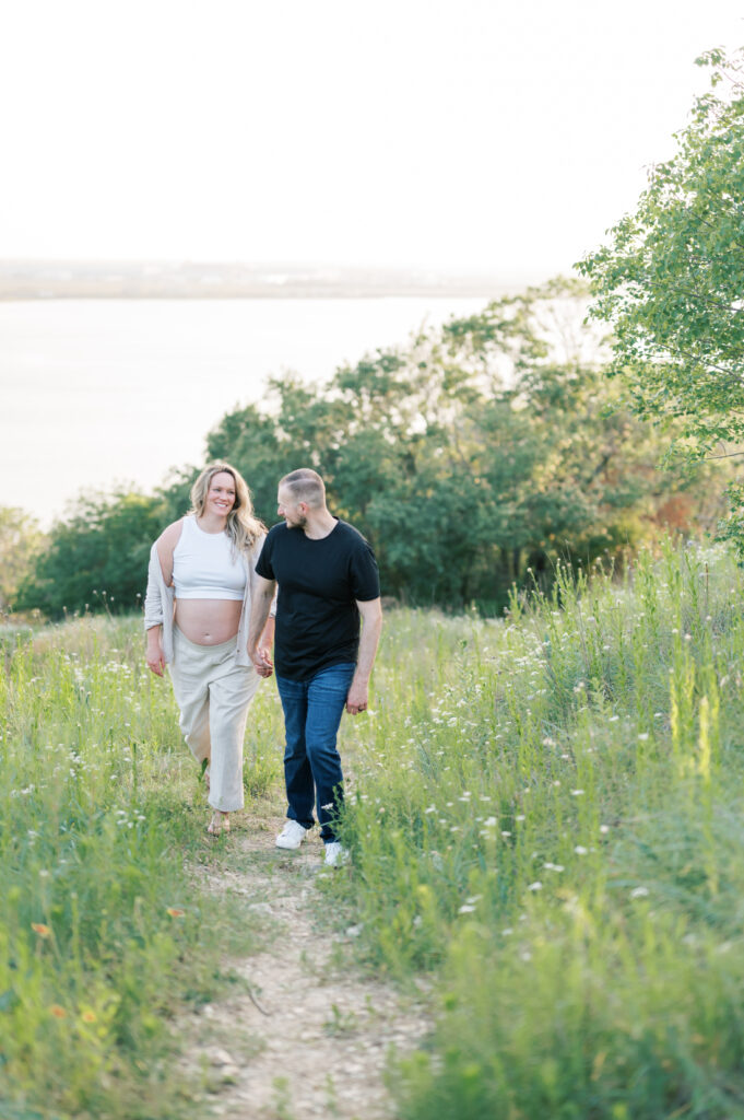 expecting parents hold hands and walk on a trail overlooking a lake