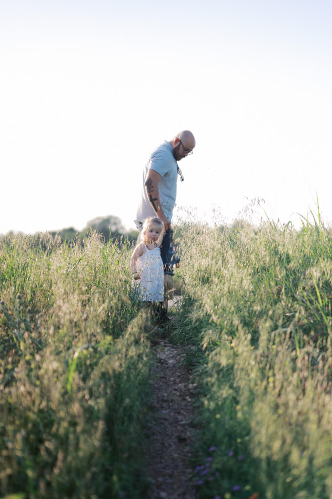 dad and toddler daughter walk through a field of tall grass