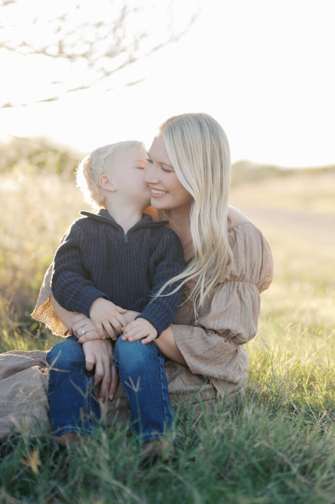 little boy kisses mom while sun shines behind them in field during fall family photos