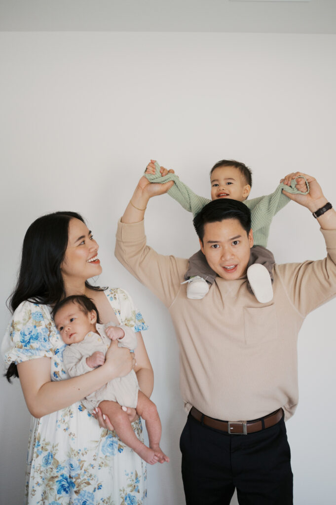 family of four gathers while mom holds newborn baby and dad carries toddler on shoulders