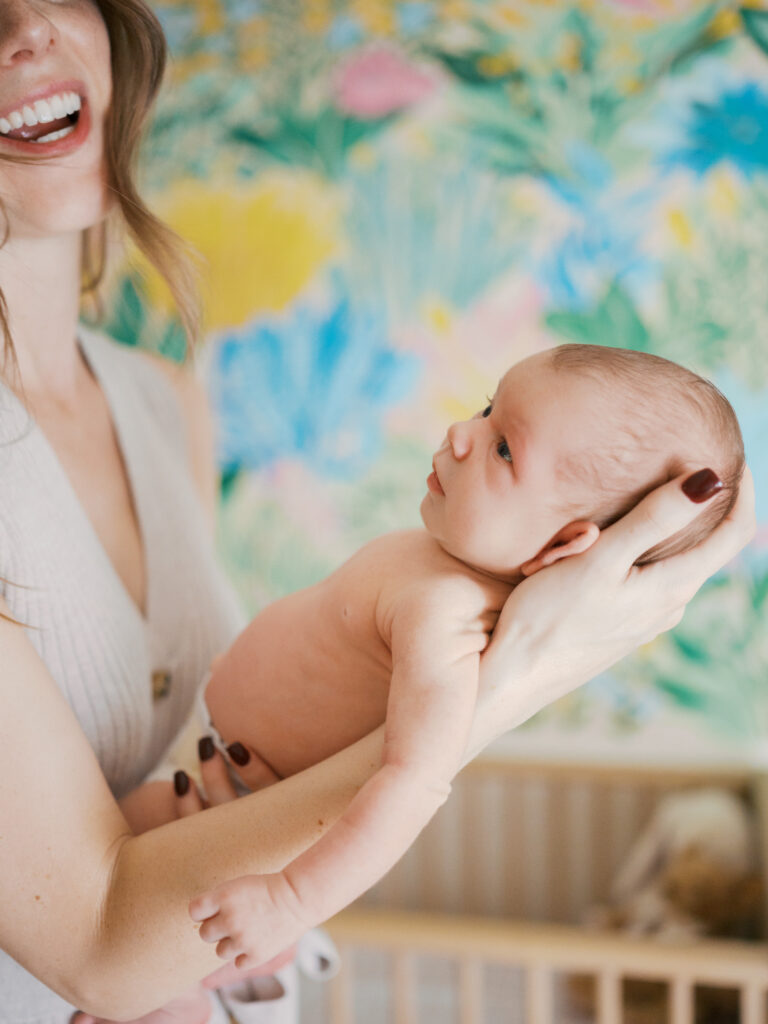 newborn baby looks up at smiling mom with nursery decorated wall behind them, in home lifestyle newborn photos in fort worth