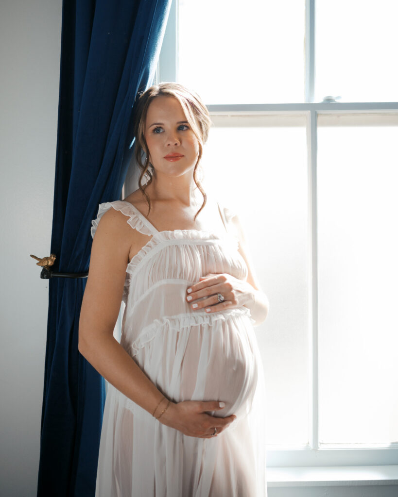 expecting mom holds baby bump next to window during maternity photos