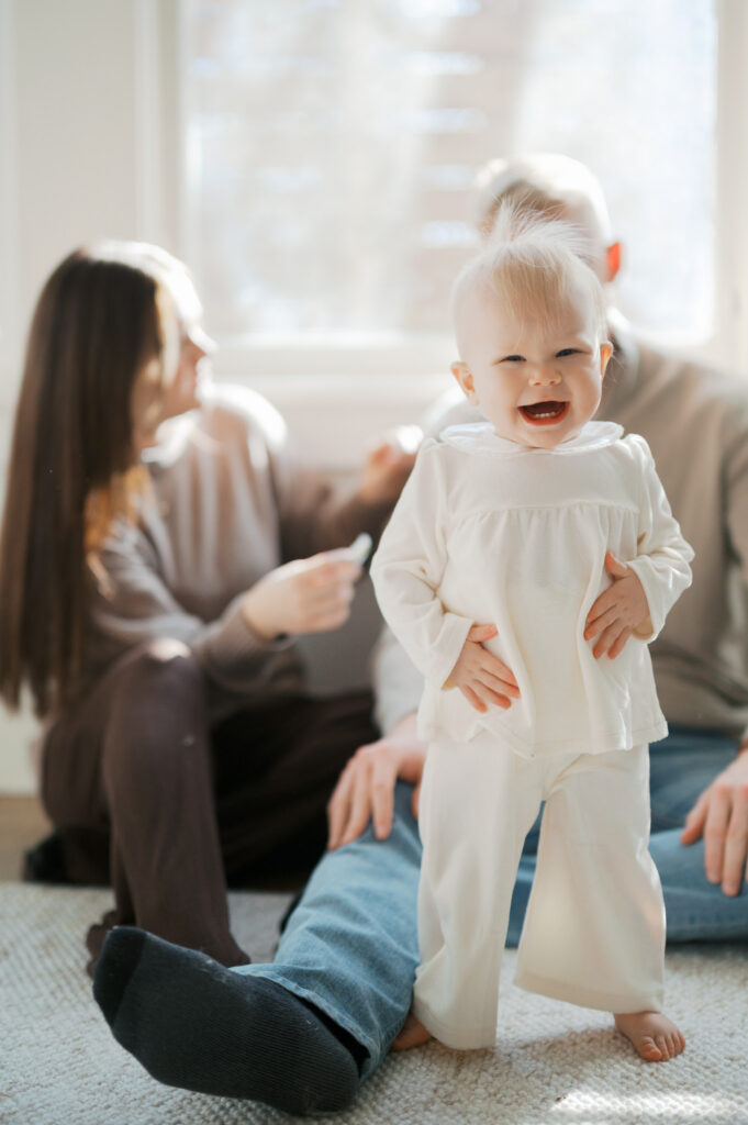one year old smiles at camera while parents in background look at each other
